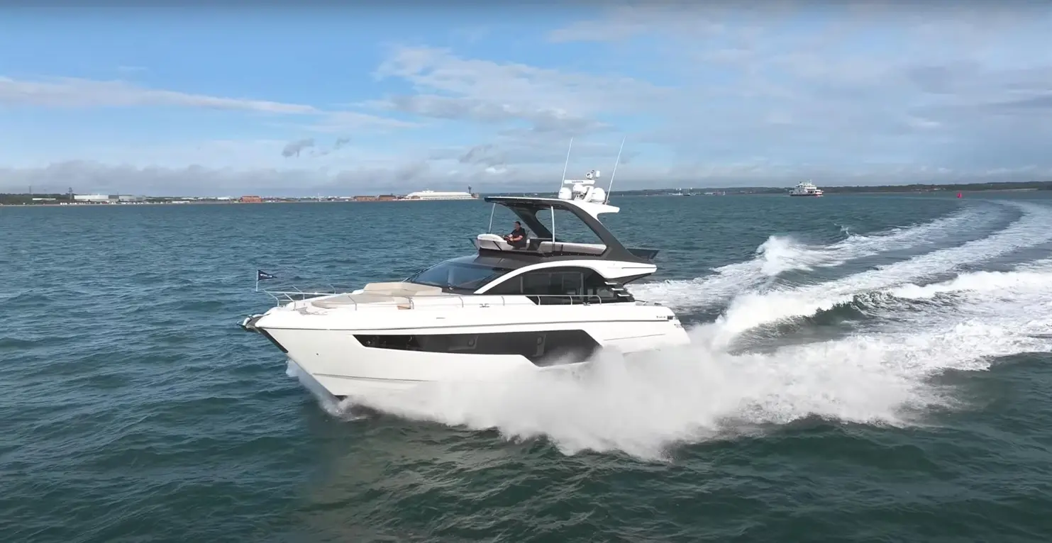 Sleek white motor yacht cutting through blue waters, with a person at the helm and coastal landscape in the background.